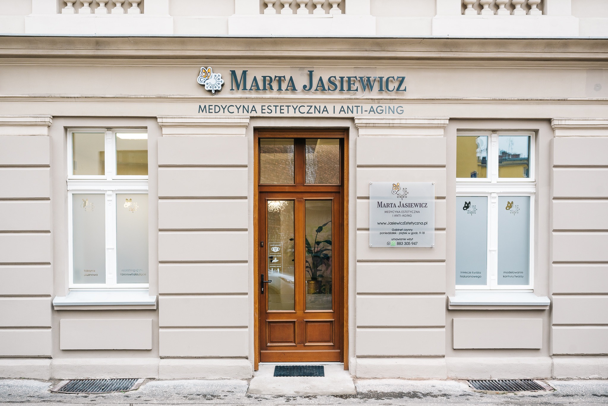 Fasada budynku z szyldem 'Marta Jasiewicz Medycyna Estetyczna i Anti-Aging', drewniane drzwi wejściowe z szybami i okna z naklejkami informującymi o zabiegach, na ścianie tabliczka informacyjna...