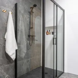 Nowoczesna kabina prysznicowa z czarną ramą, szarymi płytkami imitującymi kamień, deszczownicą w kolorze grafitowym i białym ręcznikiem na złotym wieszaku.