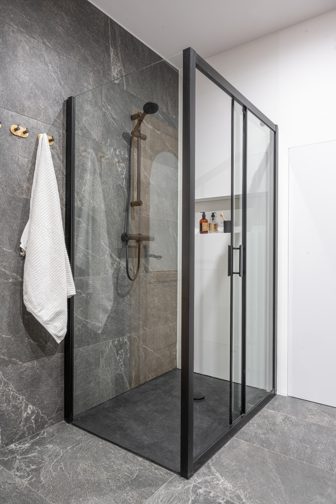 Nowoczesna kabina prysznicowa z czarną ramą, szarymi płytkami imitującymi kamień, deszczownicą w kolorze grafitowym i białym ręcznikiem na złotym wieszaku.