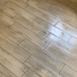 Jasne, podłużne płytki podłogowe imitujące drewno ułożone w łazience, z widocznymi fugami i odbiciami światła.