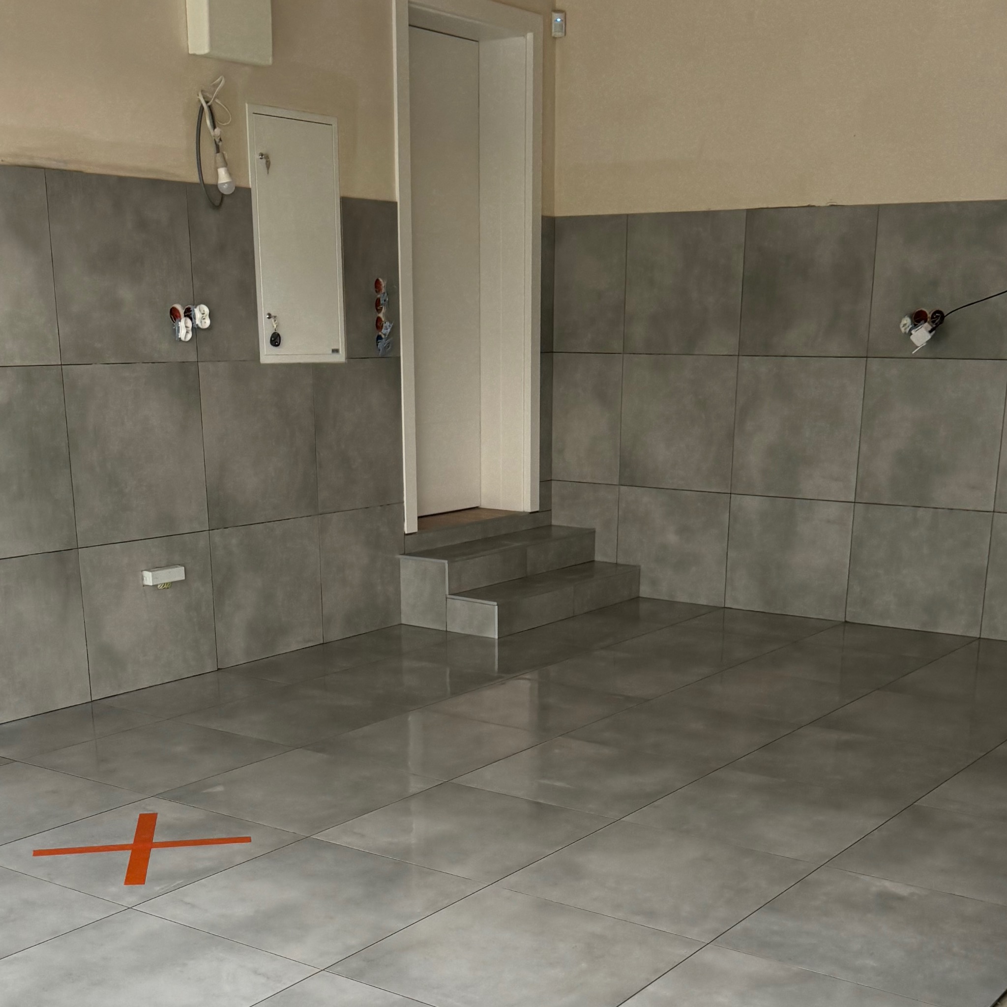 Wnętrze w trakcie wykańczania z szarymi, kwadratowymi płytkami na ścianach i podłodze, widoczne niedokończone instalacje elektryczne i hydrauliczne oraz pomarańczowy znak na podłodze.