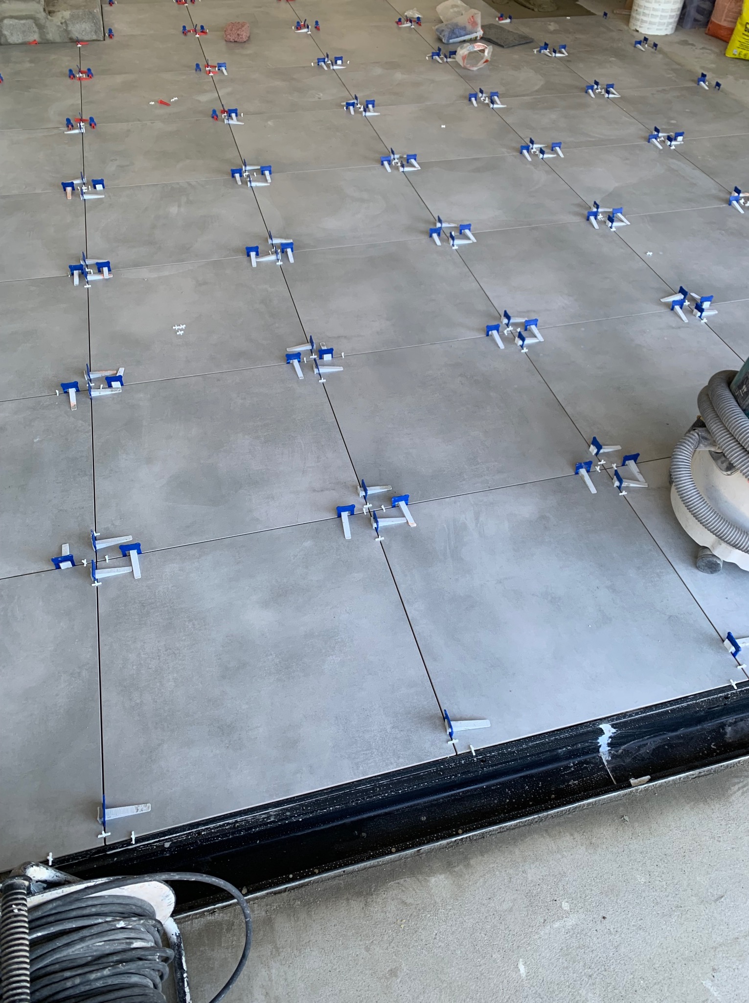 Szare płytki podłogowe w trakcie układania z użyciem systemu poziomowania płytek (klipsy i kliny), widoczny odkurzacz przemysłowy i zwinięty kabel.