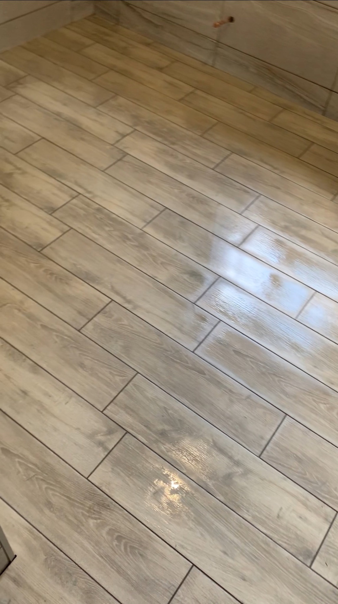 Jasne, podłużne płytki podłogowe imitujące drewno ułożone w łazience, z widocznymi fugami i odbiciami światła.