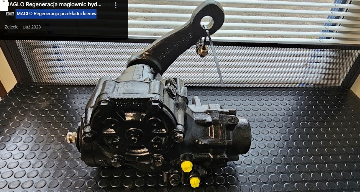 Czarna, zregenerowana przekładnia kierownicza z widocznymi żółtymi zaślepkami, umieszczona na gumowej macie z okrągłymi wypustkami; w tle siatka i żaluzje okienne.