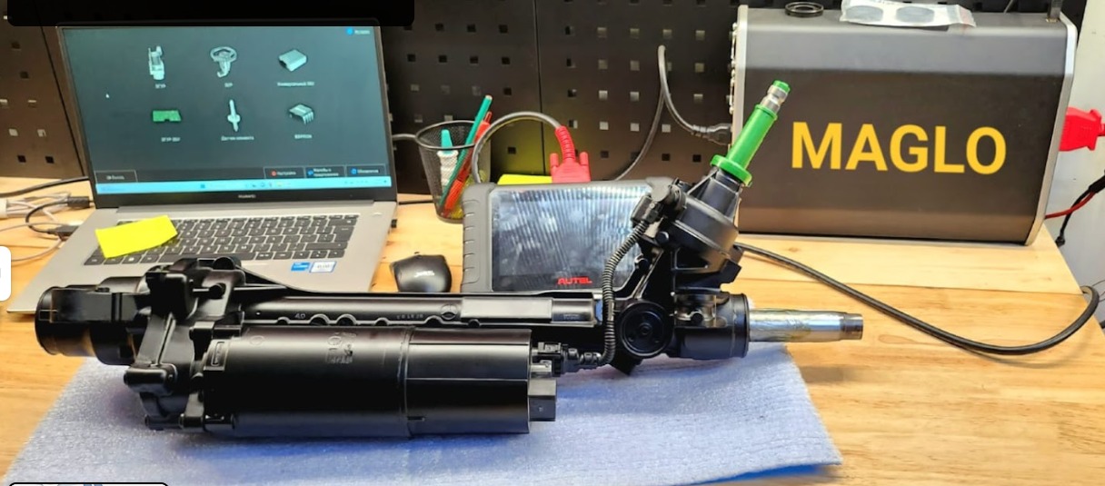 Zdemontowana, czarna przekładnia kierownicza na stole warsztatowym, podłączona do testera diagnostycznego Autel, obok laptop z oprogramowaniem diagnostycznym i skrzynka z napisem MAGLO.