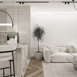 Katarzyna Wysopal - Nowoczesne wnętrze: beżowa sofa modułowa, szklany stolik, doniczkowe drzewko oliwne. Wyspa kuchenna z marmurowym blatem, białym wazonem, krzesłem barowym. Ściana z listwami, oświetlenie szynowe...