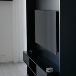 Remohouse - Minimalistyczny salon z czarną ścianką RTV, telewizorem i białymi zasłonami, widok na kuchnię z białymi meblami.