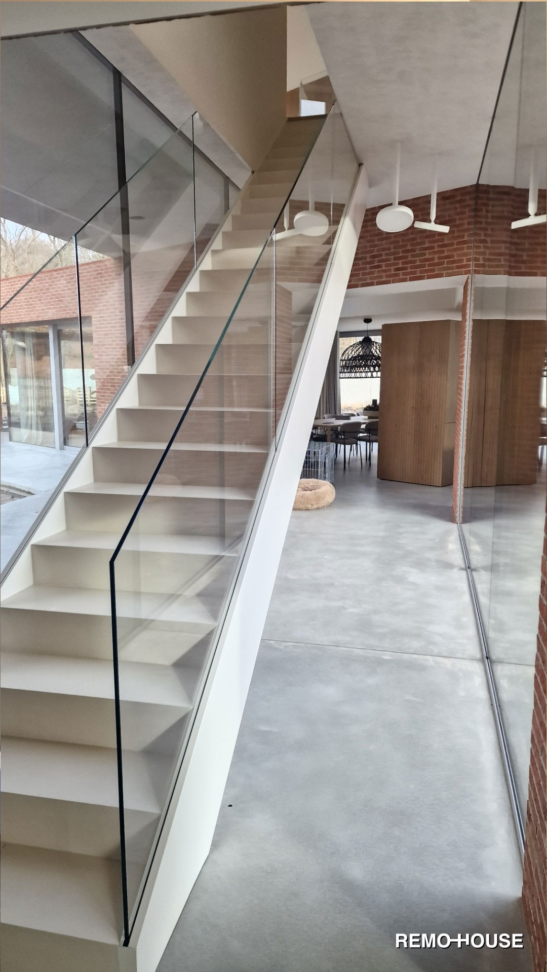 Nowoczesne schody z białymi stopniami i szklaną balustradą w przestronnym wnętrzu z betonową posadzką, widok na jadalnię z dekoracyjnym oświetleniem.