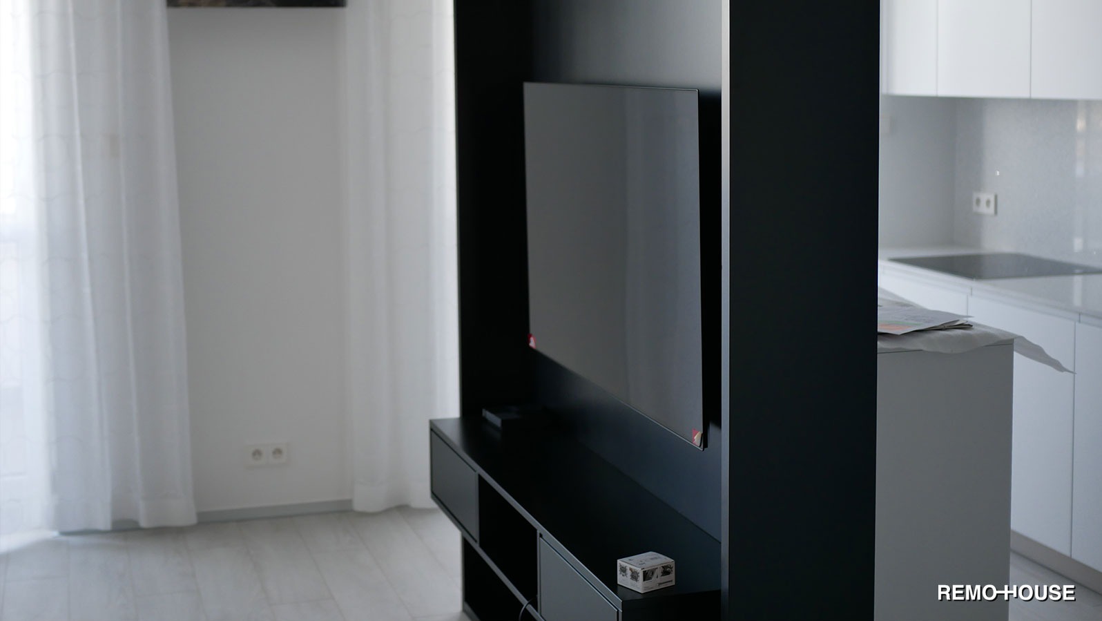 Minimalistyczny salon z czarną ścianką RTV, telewizorem i białymi zasłonami, widok na kuchnię z białymi meblami.