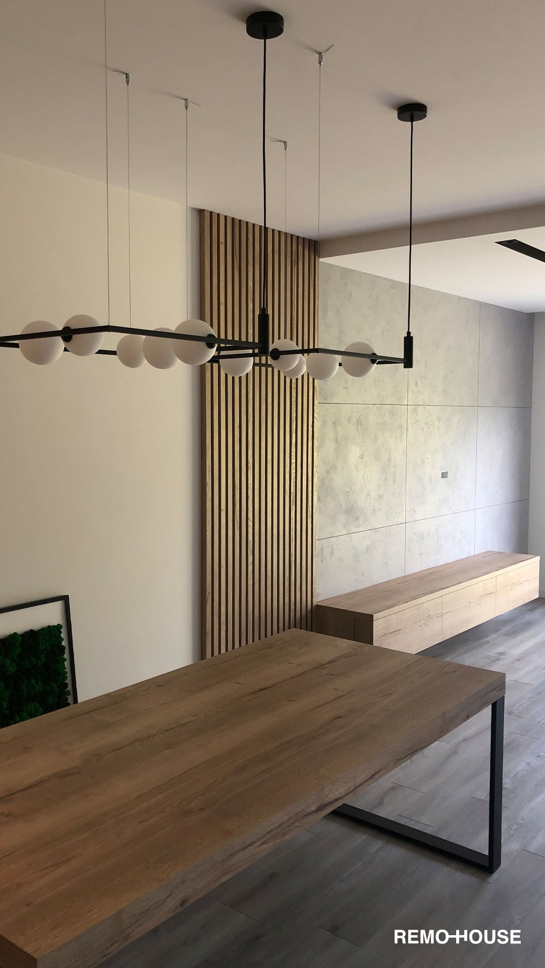 Nowoczesne wnętrze: stół z blatem z naturalnego drewna, czarna lampa sufitowa z kulistymi kloszami, dekoracyjna ściana z drewnianych listew oraz ściana z betonowych płyt, niska szafka RTV z jasnego...