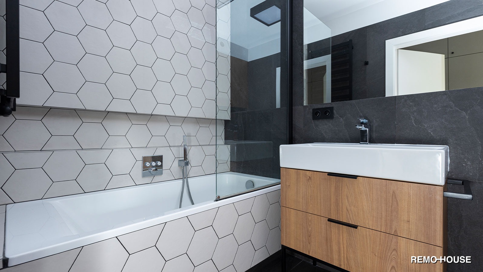Nowoczesna łazienka z wanną obudowaną heksagonalnymi płytkami, szklanym parawanem nawannowym, umywalką nablatową na drewnianej szafce i ciemnoszarą ścianą z lustrem.