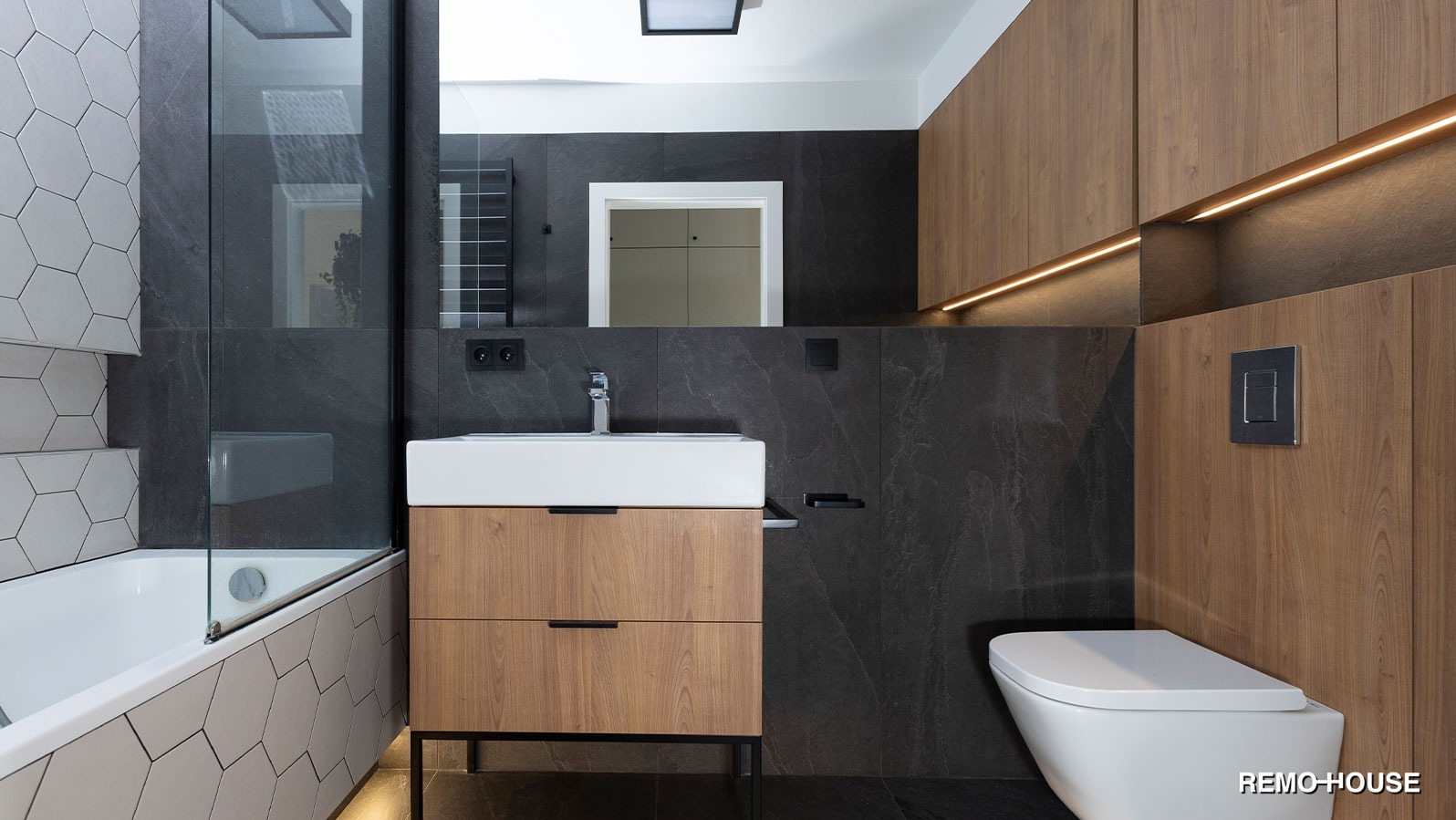 Nowoczesna łazienka z heksagonalnymi płytkami przy wannie, ciemnoszarymi płytami imitującymi kamień na ścianach, drewnianą szafką pod umywalką na czarnych nóżkach oraz podwieszaną toaletą.