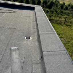 Wykończony, płaski dach pokryty papą z widocznym odpływem wody i fragmentem niezabezpieczonej krawędzi papy w słoneczny dzień.