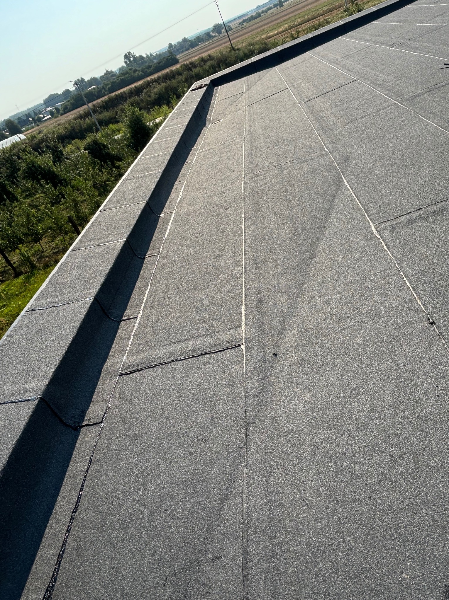 Widok z góry na płaski dach pokryty papą z widocznymi zgrzewami, na tle zieleni drzew i pól uprawnych w oddali.