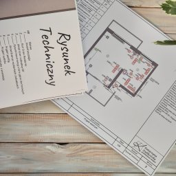 Miauart - Rysunek techniczny z elektryką wnętrza, w tym rzut ścian, rozmieszczenie gniazdek i włączników, leżący na drewnianym blacie obok spisu inwentaryzacyjnego i rośliny.