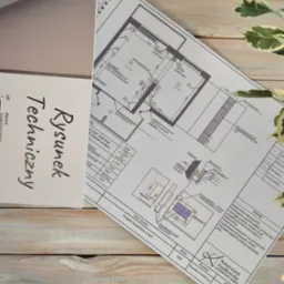 Rysunek techniczny projektu malowania ścian, z detalami wykończenia, umieszczony na drewnianym stole obok rośliny doniczkowej i notesu z napisem 'Rysunek Techniczny'.