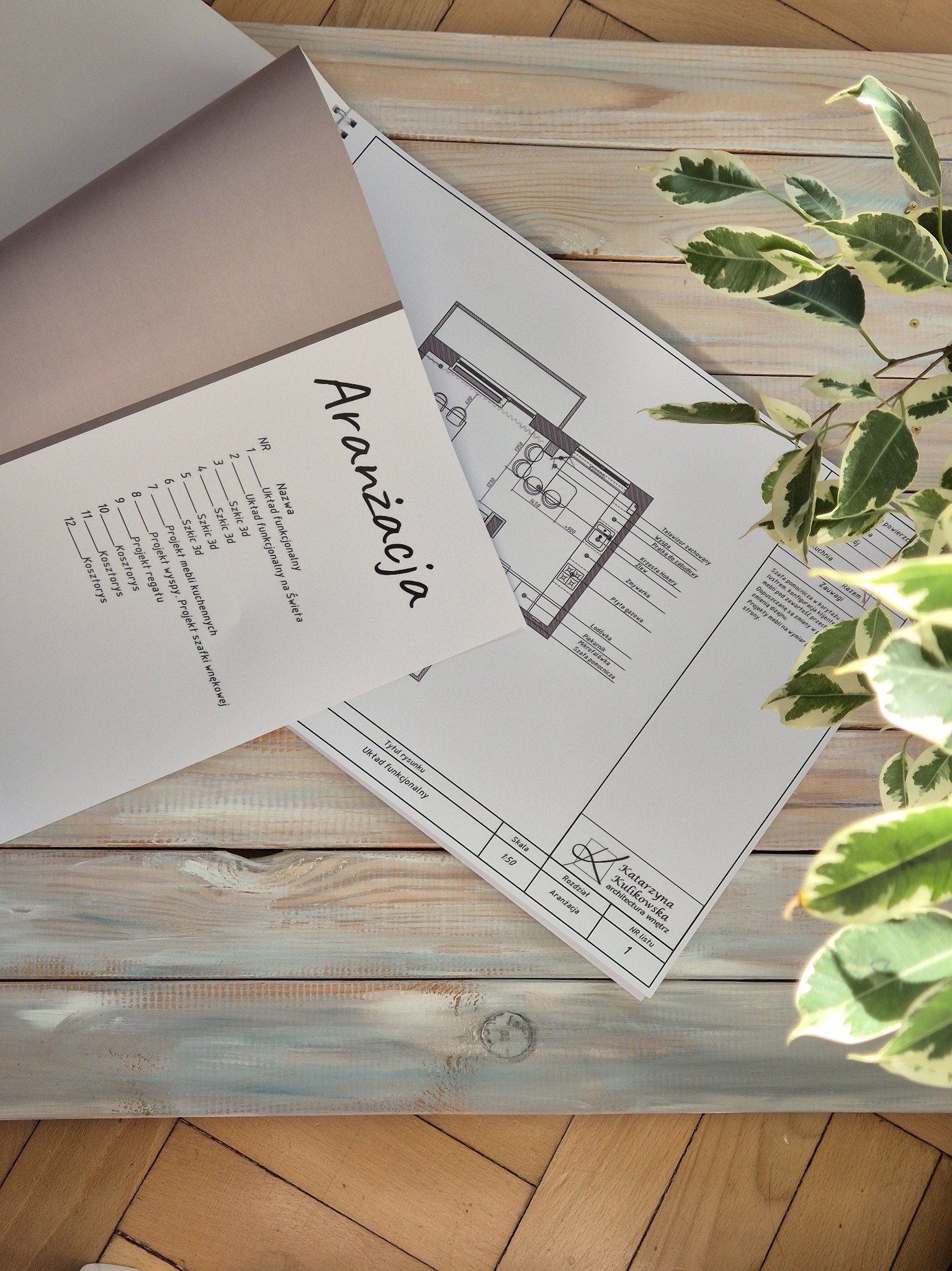 Plan układu funkcjonalnego kuchni z wyspą, fragment projektu aranżacji wnętrz, widoczny na drewnianym blacie obok rośliny doniczkowej.