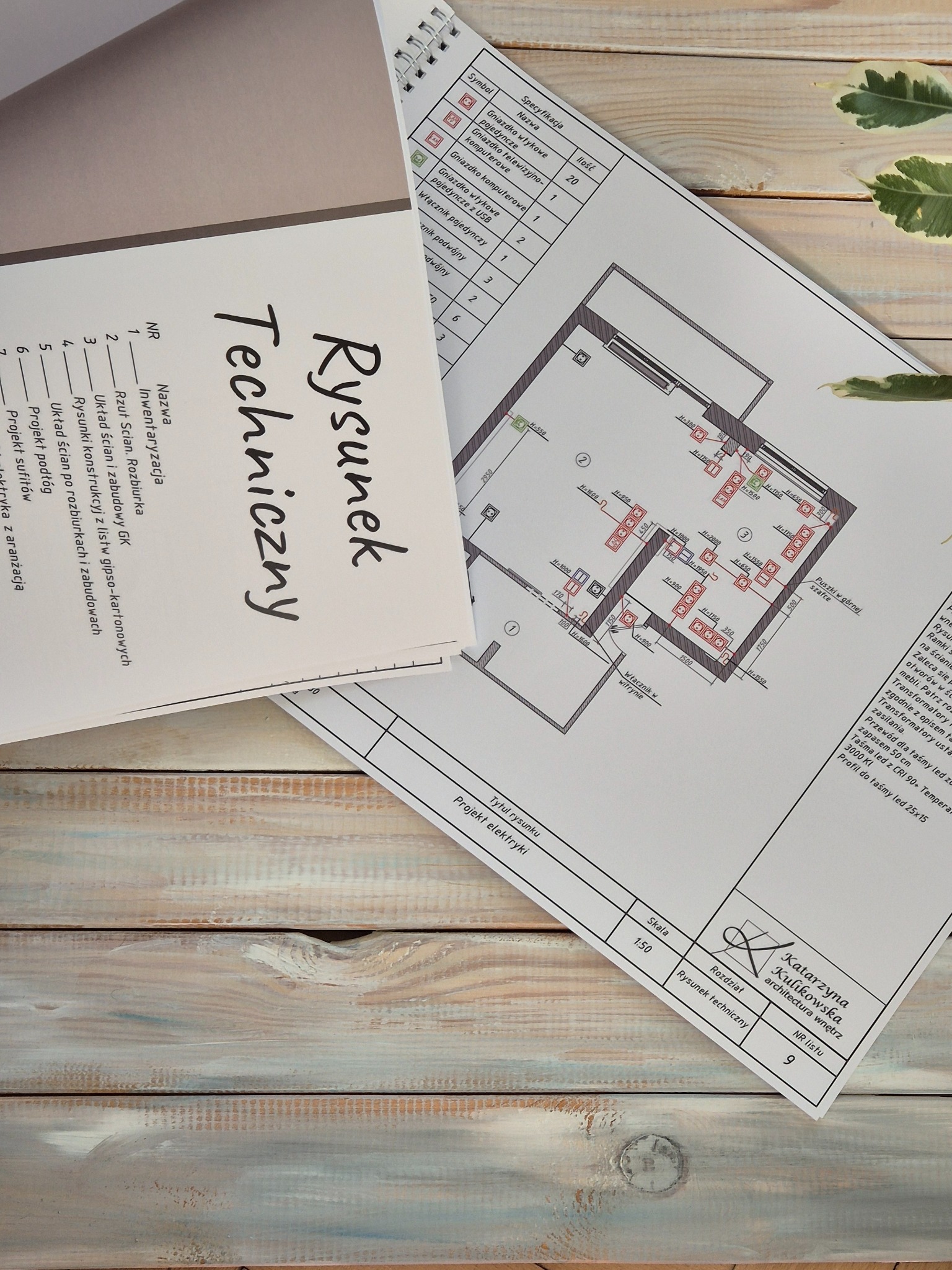 Rysunek techniczny z elektryką wnętrza, w tym rzut ścian, rozmieszczenie gniazdek i włączników, leżący na drewnianym blacie obok spisu inwentaryzacyjnego i rośliny.