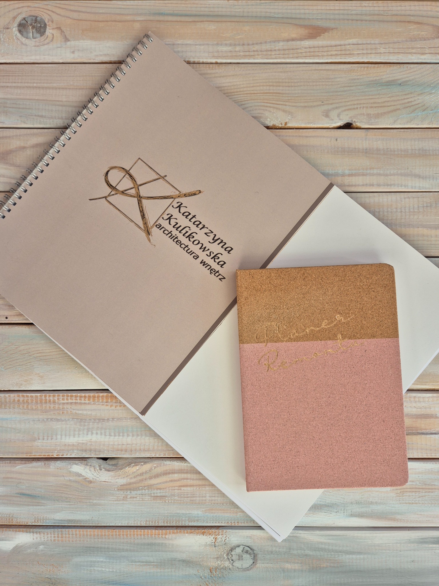 Notatnik z logo Katarzyny Kulikowskiej, architektura wnętrz, leżący na drewnianym stole obok planera w odcieniach różu i korka.
