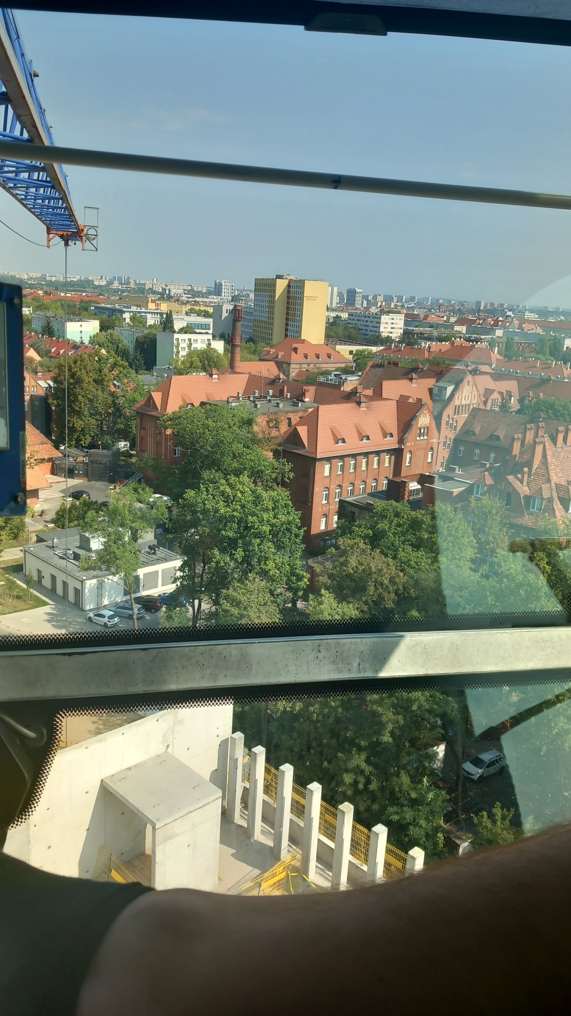 Widok z kabiny dźwigu na miasto z dominującą ceglaną zabudową i plac budowy z betonowymi elementami w dolnej części kadru.