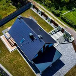 Widok z lotu ptaka na dom jednorodzinny z ciemnym, dwuspadowym pokryciem, widoczne okna dachowe, kominy, drewniana weranda z schodami, ogrodzenie oraz zagospodarowany teren wokół domu.