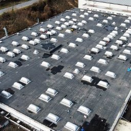 ELEMENT SPÓŁKA Z OGRANICZONĄ ODPOWIEDZIALNOŚCIĄ SPÓŁKA KOMANDYTOWA - Rozległy, płaski dach przemysłowy pokryty papą z licznymi świetlikami, widoczne ślady po wodzie i ekipa pracująca przy jednym ze świetlików, ujęcie z drona.