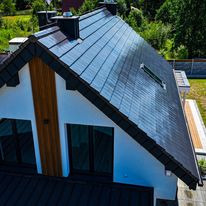 Nowoczesny dom z ciemnoszarą dachówką modułową, widoczne okno dachowe i komin, w tle zieleń drzew i fragment ogrodzenia.