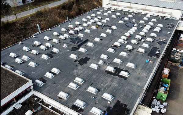 Rozległy, płaski dach przemysłowy pokryty papą z licznymi świetlikami, widoczne ślady po wodzie i ekipa pracująca przy jednym ze świetlików, ujęcie z drona.