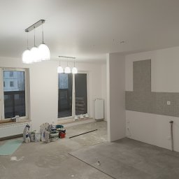 Kamil Siedlecki INDYGO - Wnętrze mieszkania w trakcie remontu: widoczne worki z gładzią i farbami przy grzejniku pod oknem, świeżo położone płytki podłogowe i fragment ściany z mozaiką w kuchni, nowe lampy sufitowe.