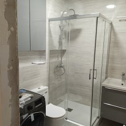 Kamil Siedlecki INDYGO - Jasna łazienka w trakcie urządzania, widoczna kabina prysznicowa, umywalka z szafką, toaleta oraz pralka z elementami montażowymi na górze, ściany wyłożone płytkami imitującymi drewno, fragment...