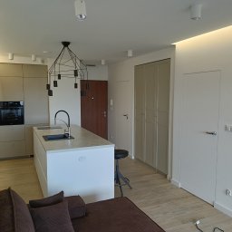 Kamil Siedlecki INDYGO - Nowoczesne wnętrze mieszkania w Białymstoku z wyspą kuchenną, minimalistycznym oświetleniem i brązową sofą w tle.