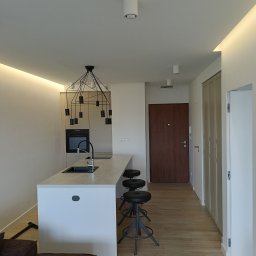 Kamil Siedlecki INDYGO - Nowoczesna kuchnia z wyspą, czarne hokery, designerska lampa wisząca nad blatem, podłoga z paneli imitujących drewno, minimalistyczne oświetlenie sufitowe.