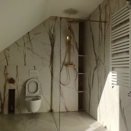 Kamil Siedlecki INDYGO - Nowoczesna łazienka na poddaszu z marmurowymi płytkami, złotą armaturą prysznicową i grzejnikiem drabinkowym, widoczny otwarty sedes wiszący.