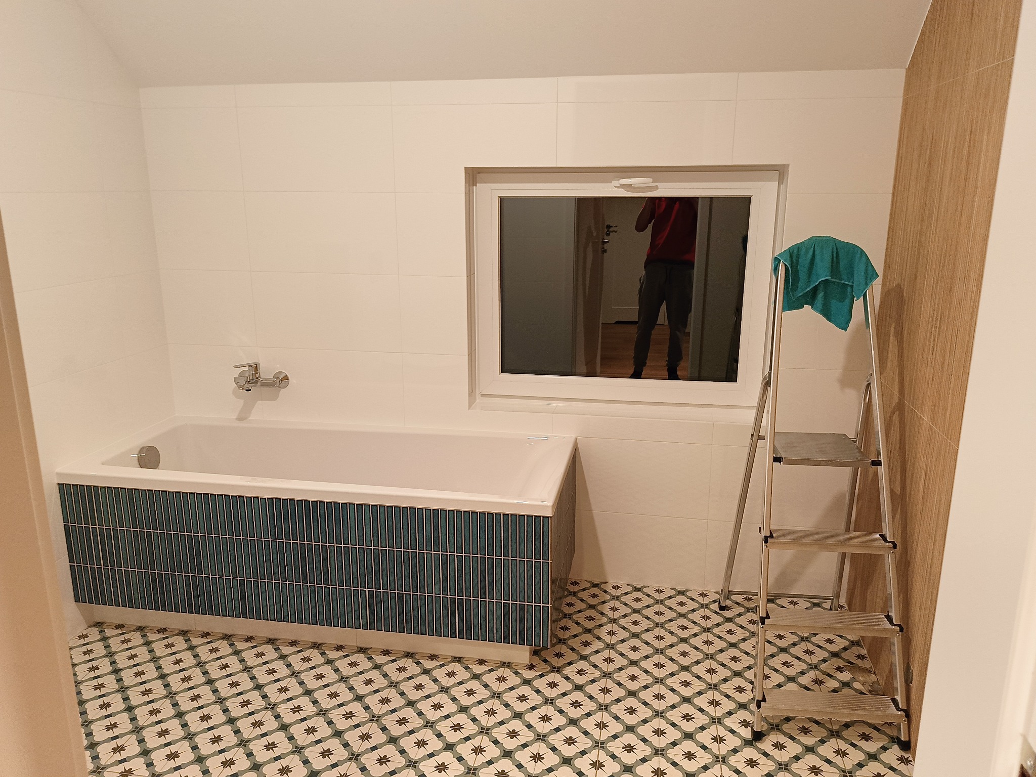 Biała wanna z mozaikową obudową w odcieniach zieleni i błękitu, na podłodze wzorzyste płytki, drabina z zielonym ręcznikiem w tle, okno z odbiciem osoby w środku.