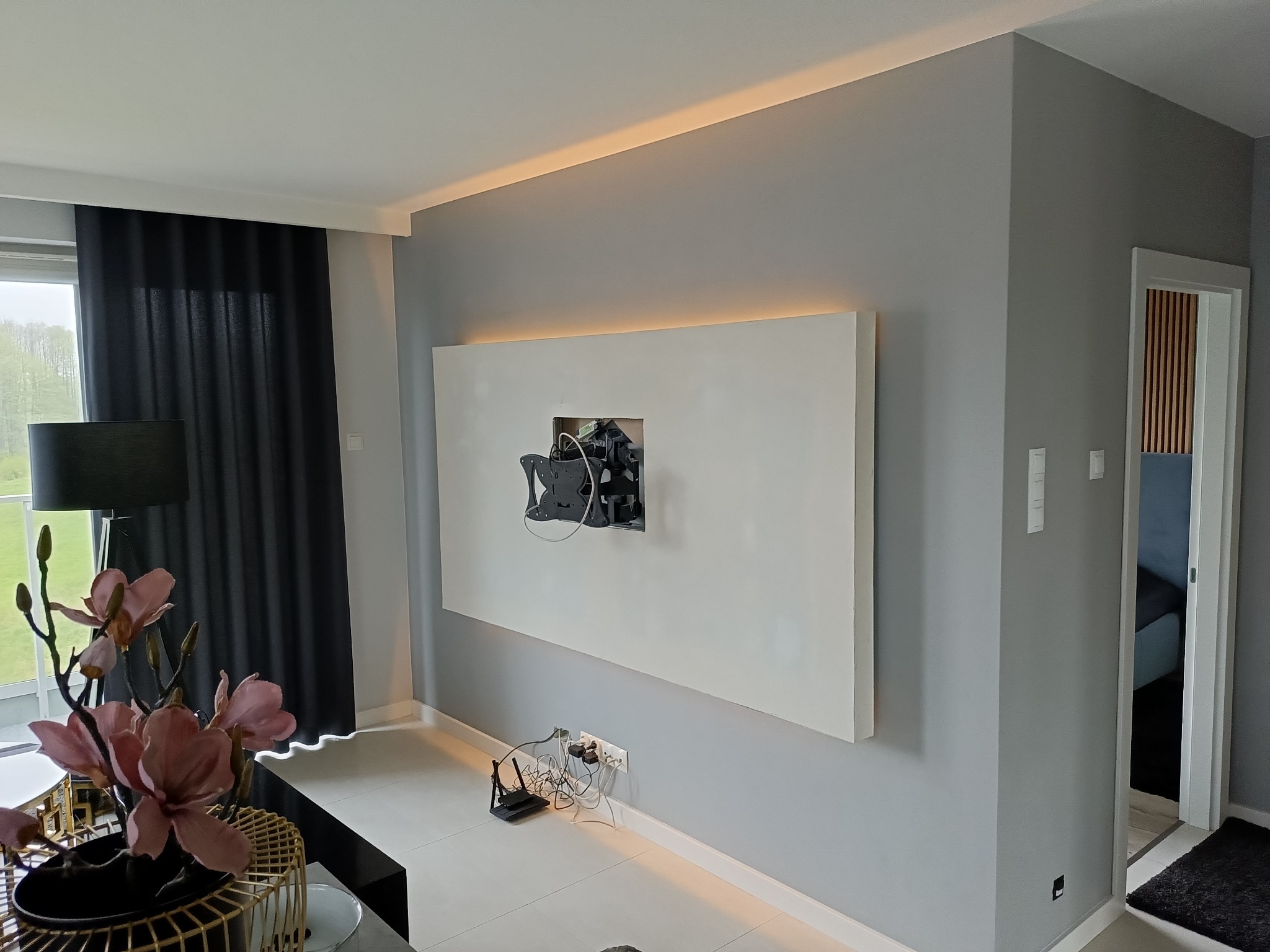 Ściana w salonie z przygotowanym miejscem na telewizor, widoczne kable i gniazdka, oświetlenie LED nad panelem, dekoracyjne kwiaty w rogu pokoju.