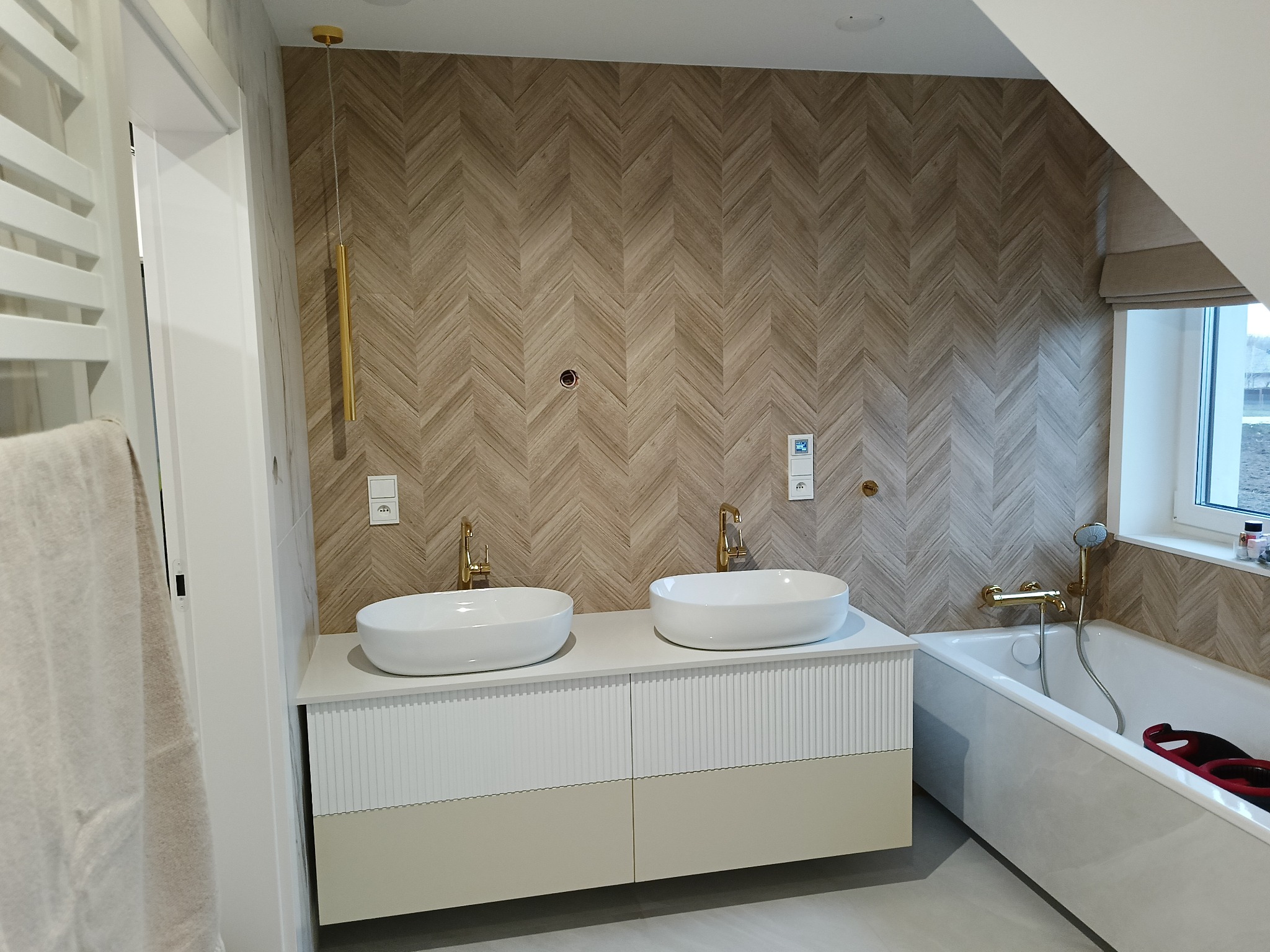 Nowoczesna łazienka na poddaszu z drewnianą ścianą w jodełkę, podwójną umywalką nablatową na szafce z frezowanymi frontami, złotą armaturą i białą wanną.