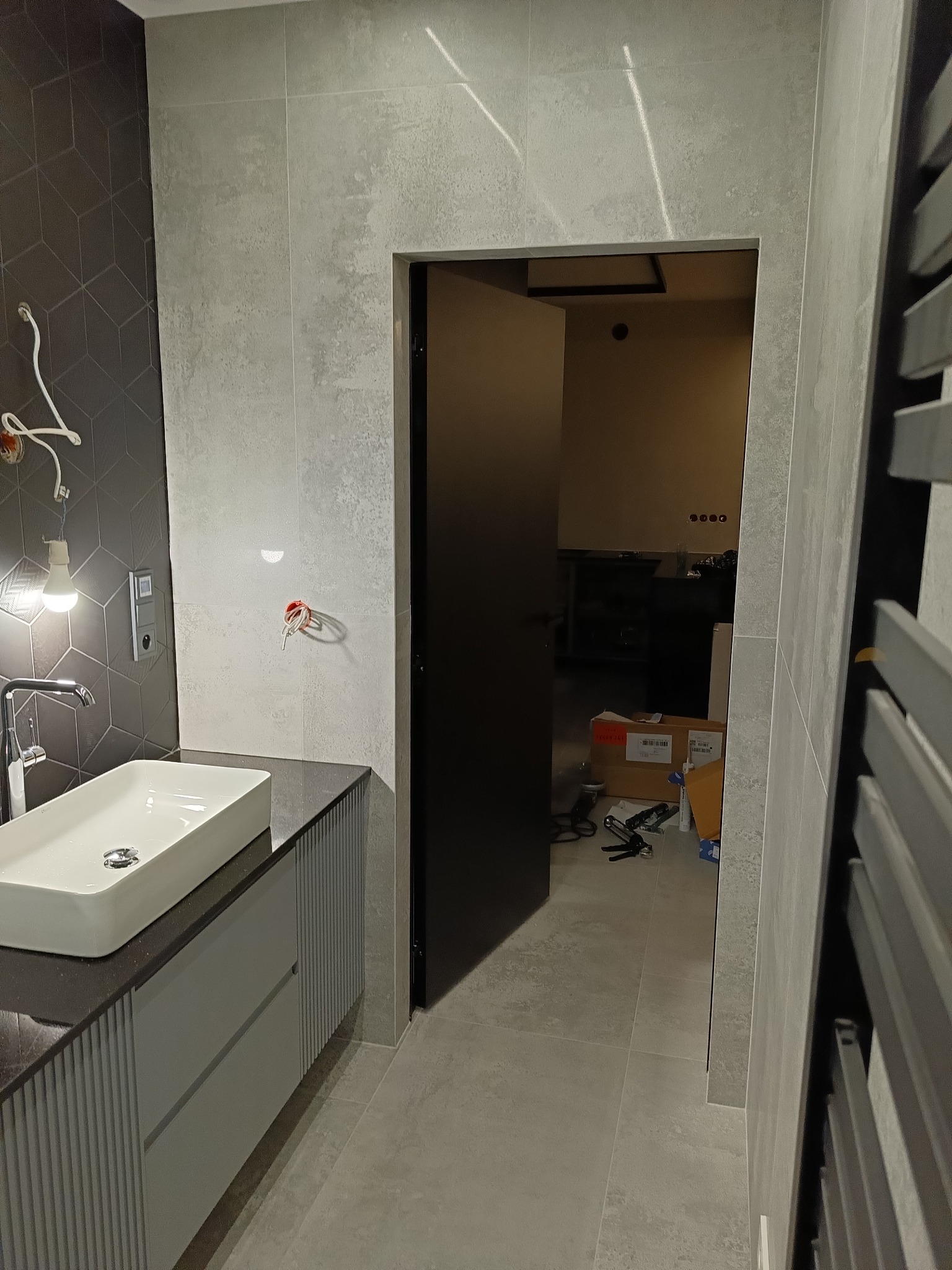 Nowoczesna łazienka w szarościach z białą umywalką nablatową, widoczne niedokończone podłączenia elektryczne i narzędzia w przejściu do innego pomieszczenia.