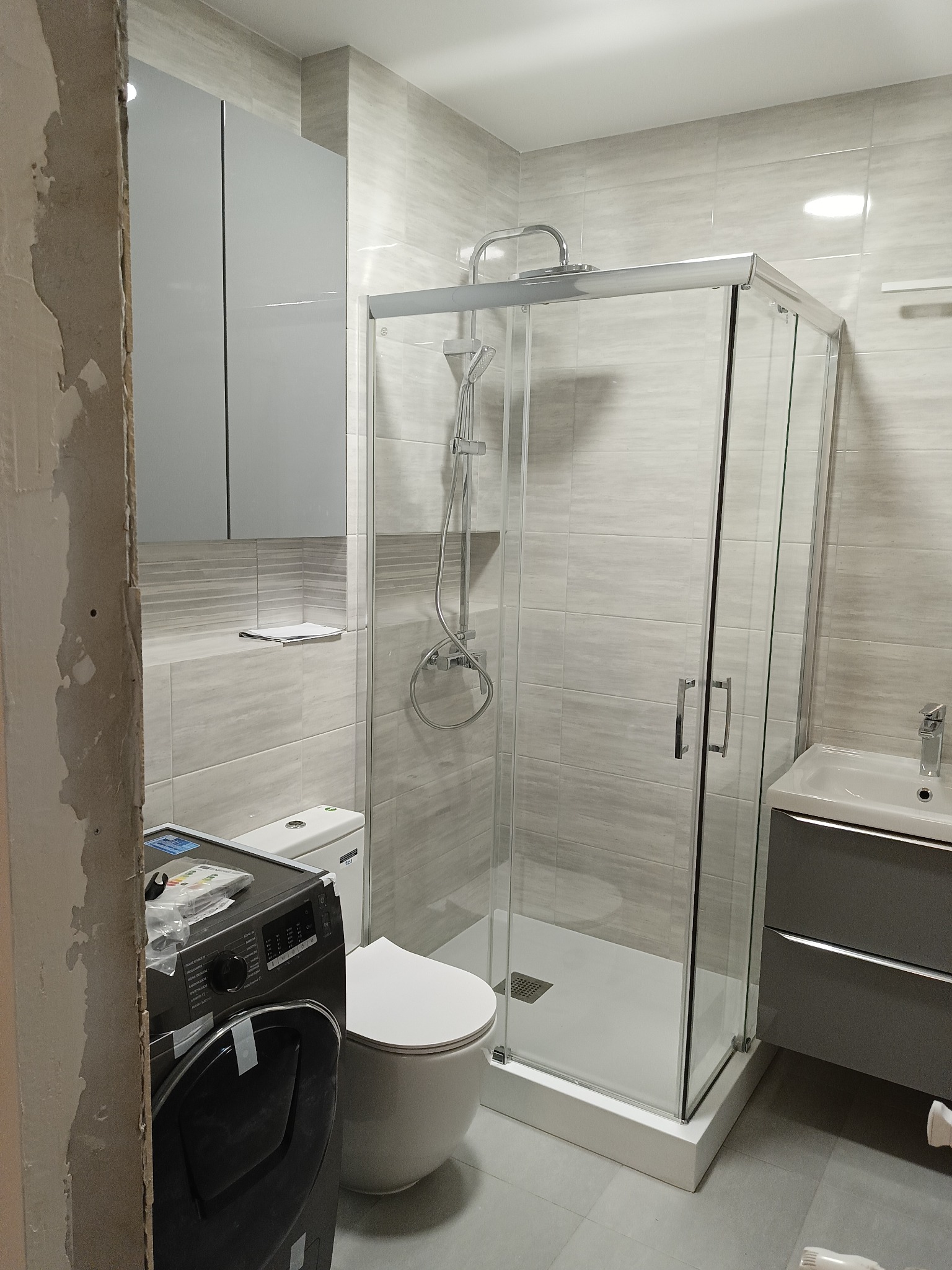 Jasna łazienka w trakcie urządzania, widoczna kabina prysznicowa, umywalka z szafką, toaleta oraz pralka z elementami montażowymi na górze, ściany wyłożone płytkami imitującymi drewno, fragment...