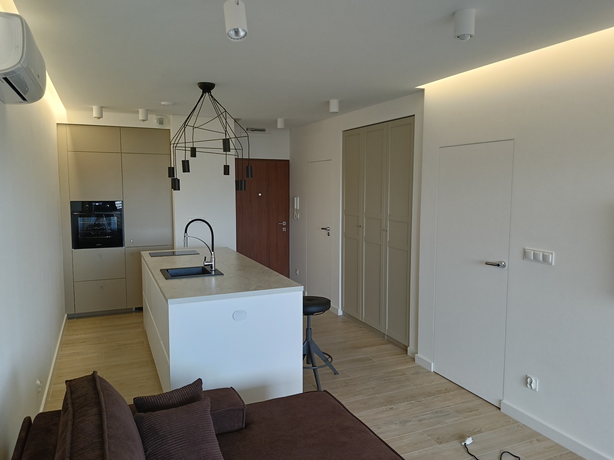 Nowoczesne wnętrze mieszkania w Białymstoku z wyspą kuchenną, minimalistycznym oświetleniem i brązową sofą w tle.