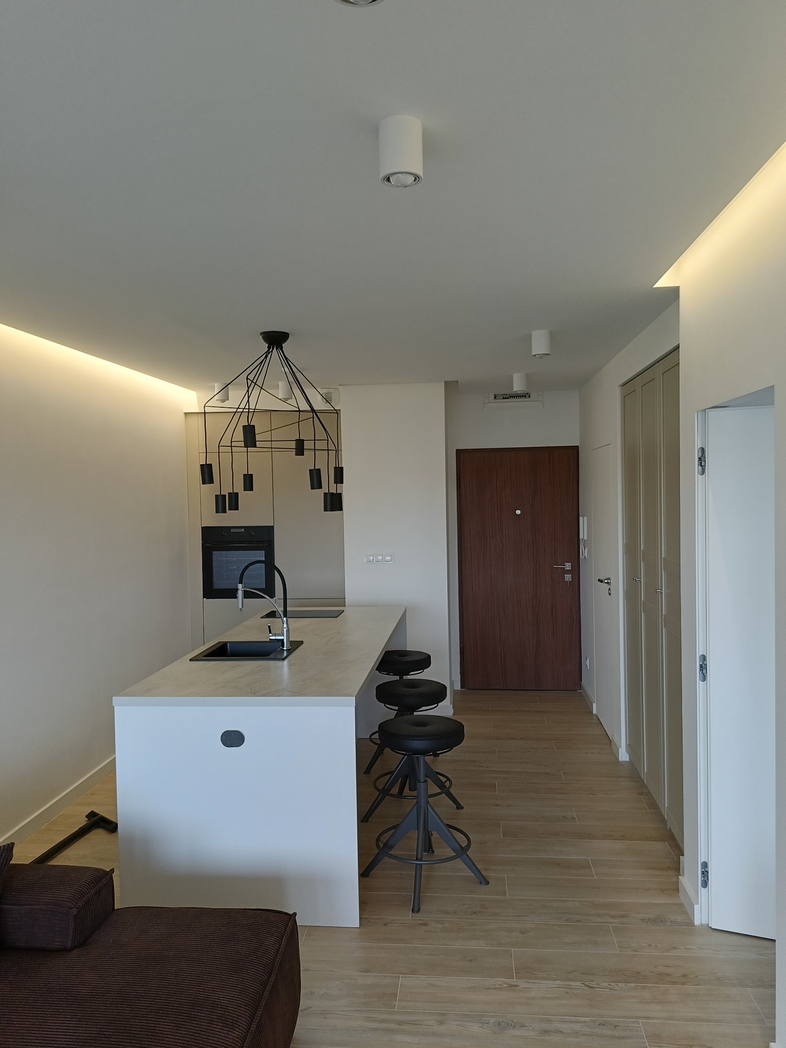 Nowoczesna kuchnia z wyspą, czarne hokery, designerska lampa wisząca nad blatem, podłoga z paneli imitujących drewno, minimalistyczne oświetlenie sufitowe.