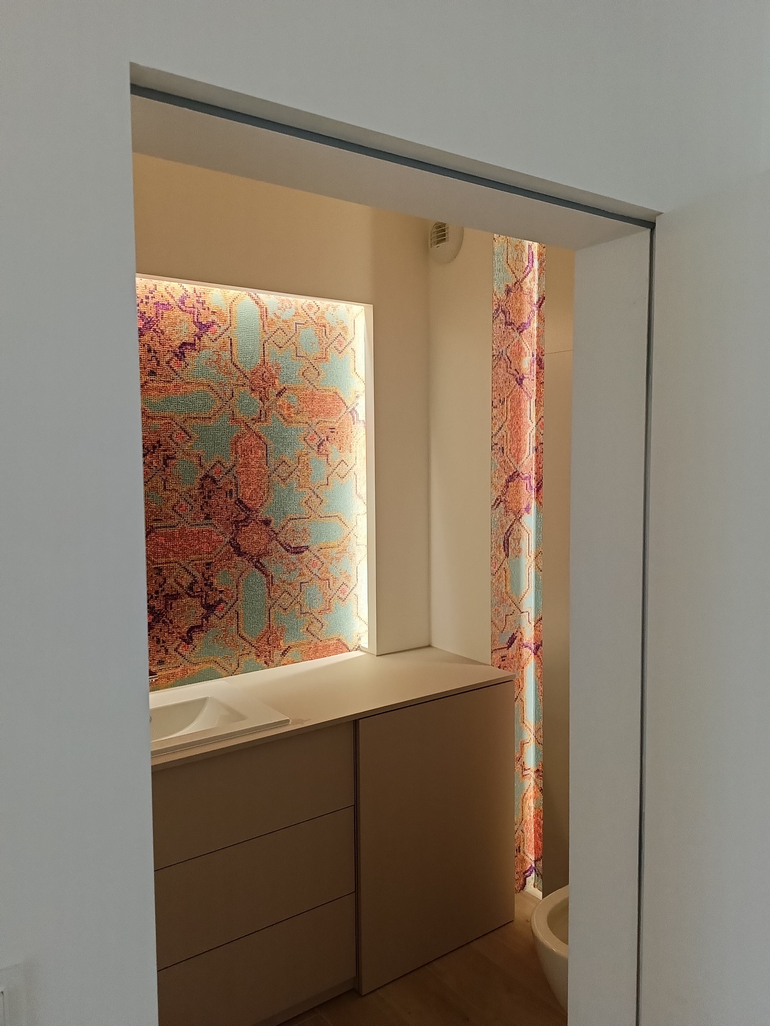 Nowoczesna łazienka z podświetlanym panelem dekoracyjnym o wzorze orientalnym, umywalką wpuszczaną w szafkę z szufladami i fragmentem toalety widocznym w tle.