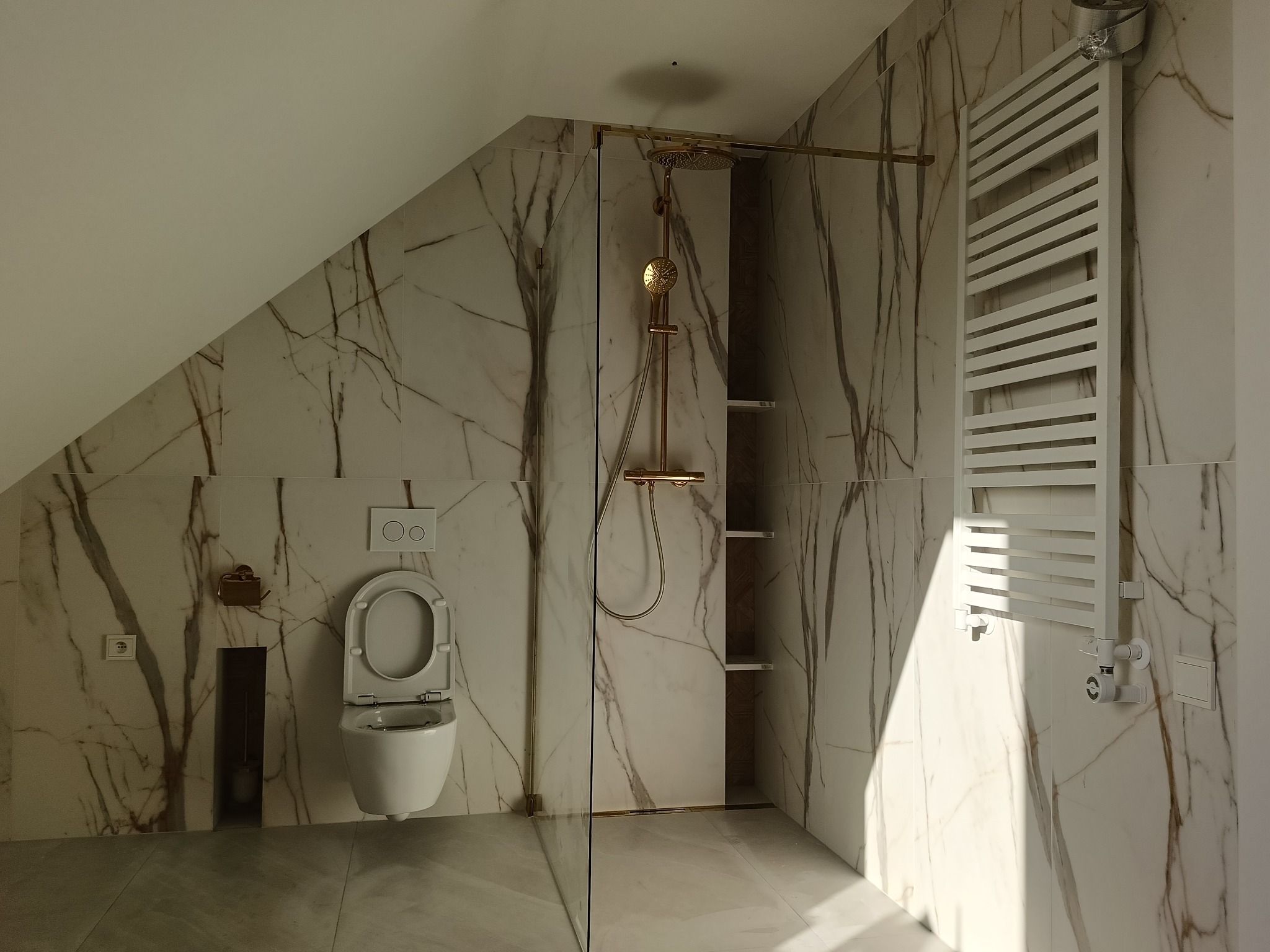 Nowoczesna łazienka na poddaszu z marmurowymi płytkami, złotą armaturą prysznicową i grzejnikiem drabinkowym, widoczny otwarty sedes wiszący.