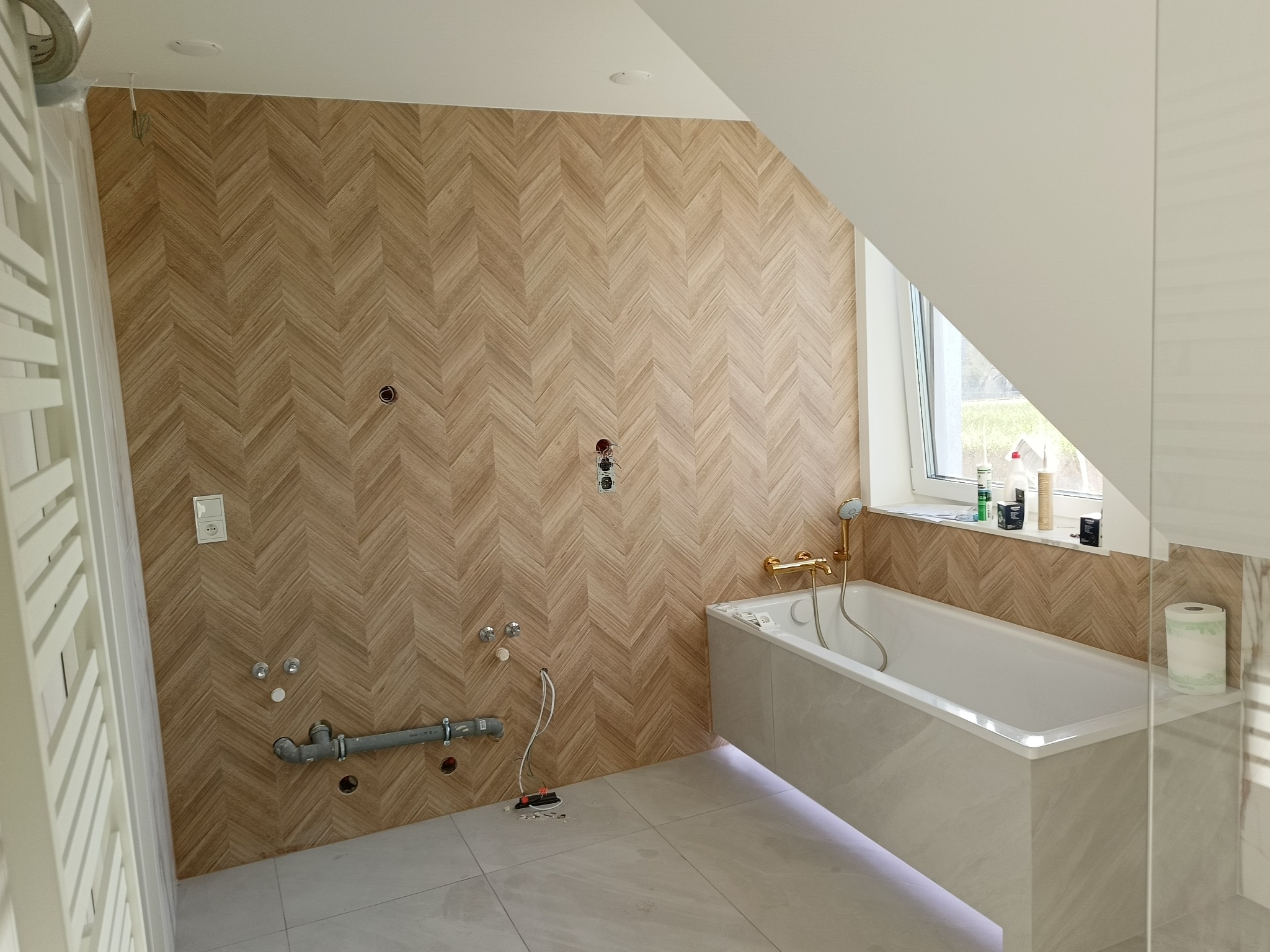 Łazienka w trakcie remontu: drewnopodobna ściana w jodełkę, biała wanna z złotą armaturą, przygotowane instalacje wodno-kanalizacyjne, grzejnik drabinkowy z boku.