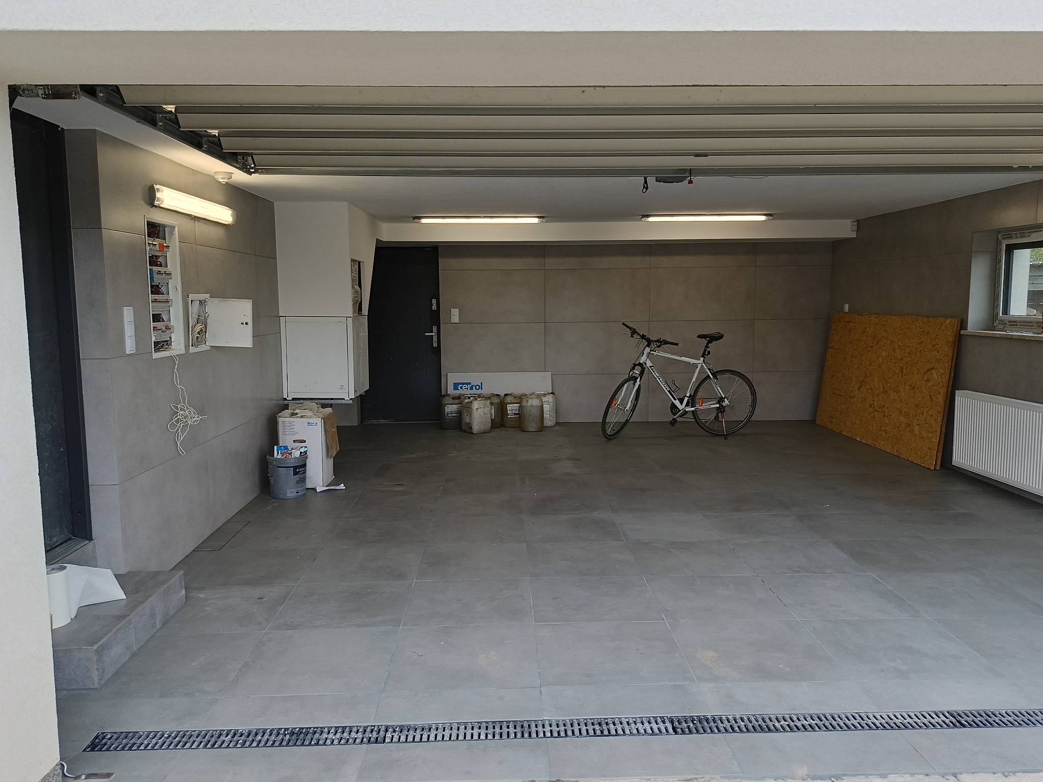 Wykończony garaż z szarymi płytkami na ścianach i podłodze, rowerem, skrzynką elektryczną i kanistrami z płynami.
