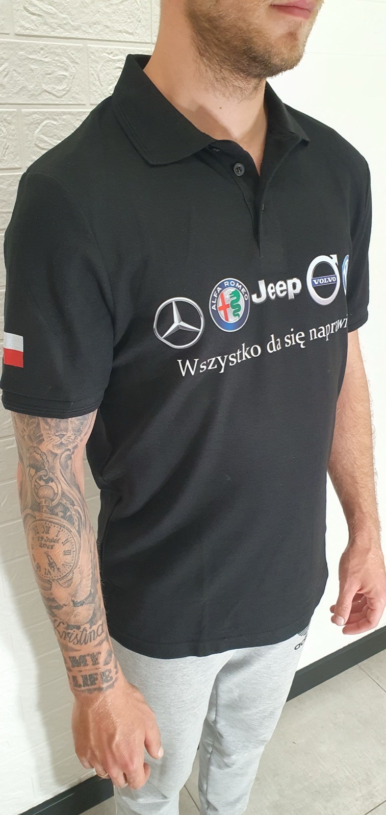 Mężczyzna w czarnej koszulce polo z nadrukowanymi logo marek samochodowych i napisem 'Wszystko da się naprawić', z tatuażem na lewym ramieniu i flagą Polski na prawym rękawie.