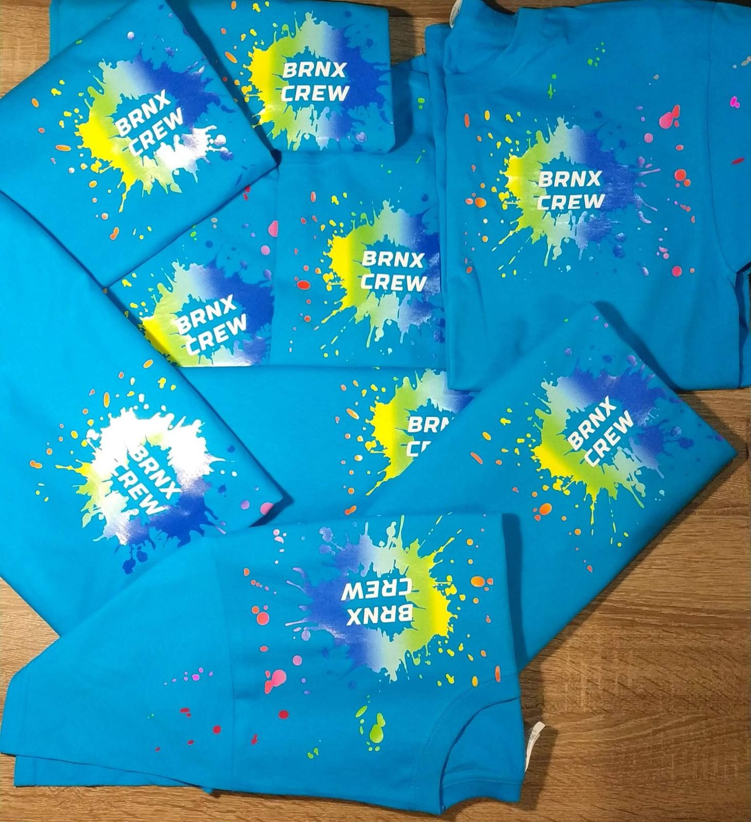 Stos turkusowych koszulek z nadrukiem 'BRNX CREW' i kolorowymi plamami farby w odcieniach żółci, bieli i niebieskiego, ułożonych na drewnianym blacie.