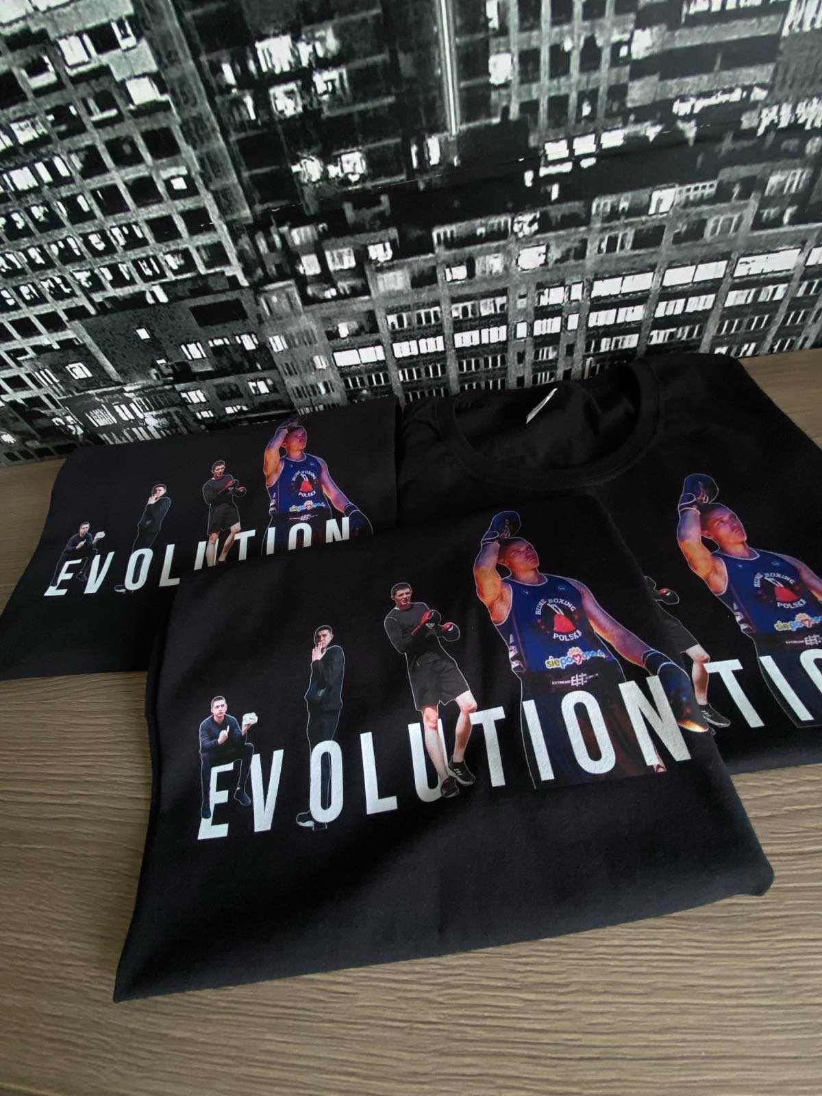Czarne koszulki z nadrukiem przedstawiającym ewolucję boksera, ułożone na drewnianym blacie na tle tapety z motywem miejskim.