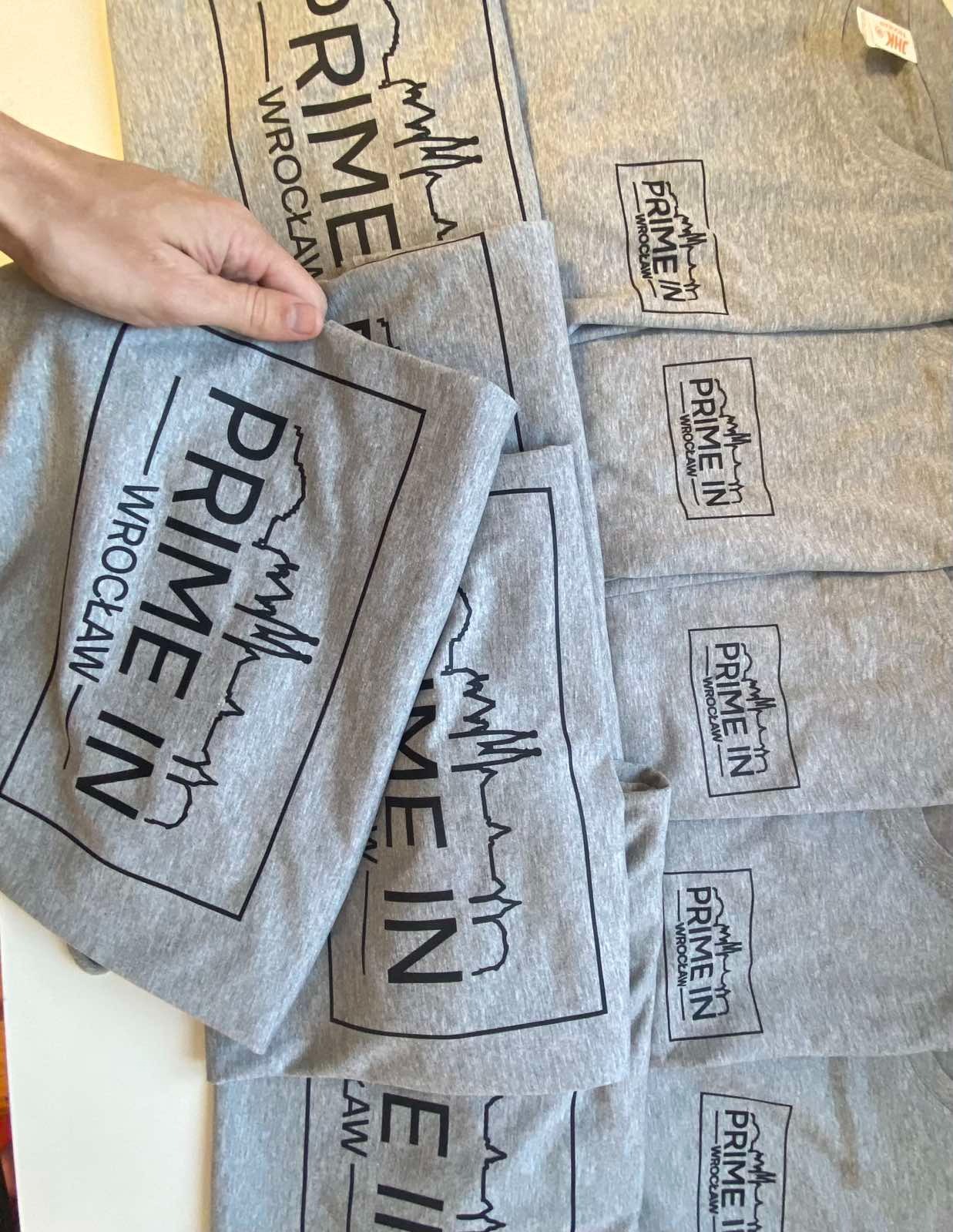 Stos szarych koszulek z nadrukowanym czarnym logo 'PRIME IN Wrocław', trzymany przez dłoń, widoczne detale nadruku i faktura materiału.