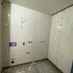 Alina Oleksiuk VIRTUS - Łazienka w trakcie wykańczania: białe płytki imitujące marmur na ścianach, widoczne otwory na instalacje, zielone krzyżyki dystansowe między płytkami, surowa betonowa podłoga z resztkami zaprawy.