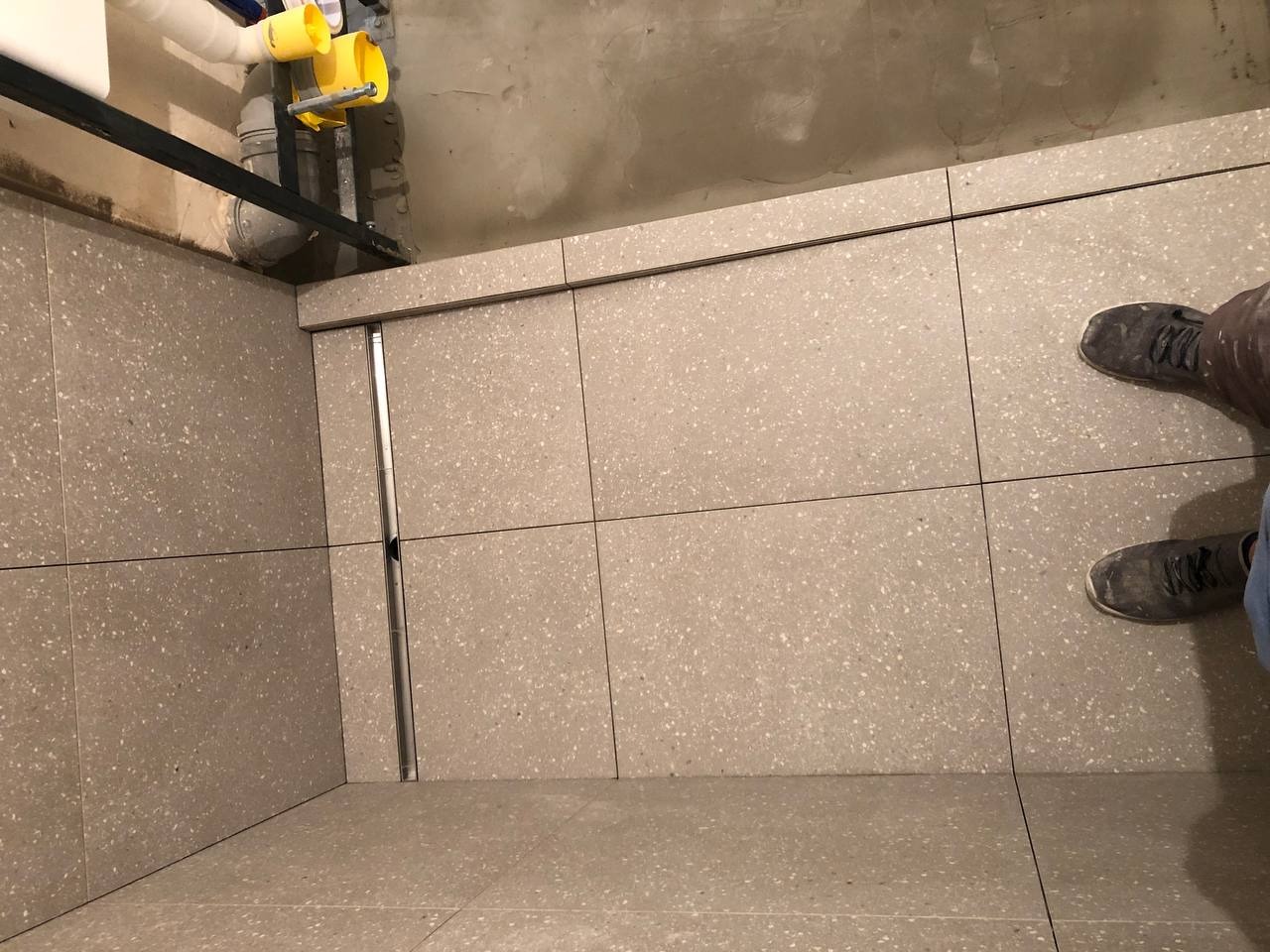 Świeżo ułożone, szare, kwadratowe płytki podłogowe z drobnym wzorem w łazience, widoczny odpływ liniowy i fragment instalacji sanitarnej pod sufitem, robocze buty glazurnika w prawym górnym rogu.